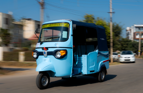 Light blue electric three-wheeler tuk-tuk driving on an Indian suburban road, a compact last-mile transport option