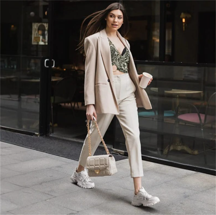 Woman in smart beige blazer and tailored trousers pairing formal workwear with chunky white casual sneakers on city sidewalk