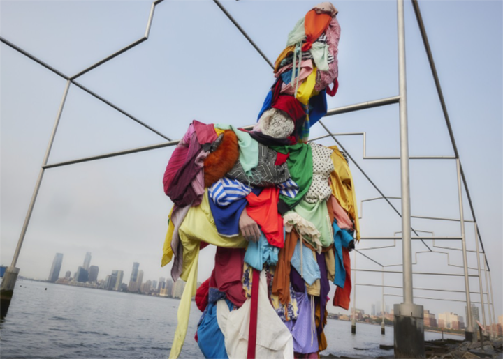 Art installation figure covered entirely in piled secondhand clothing, overlooking city waterfront to visualize global surplus of donated apparel